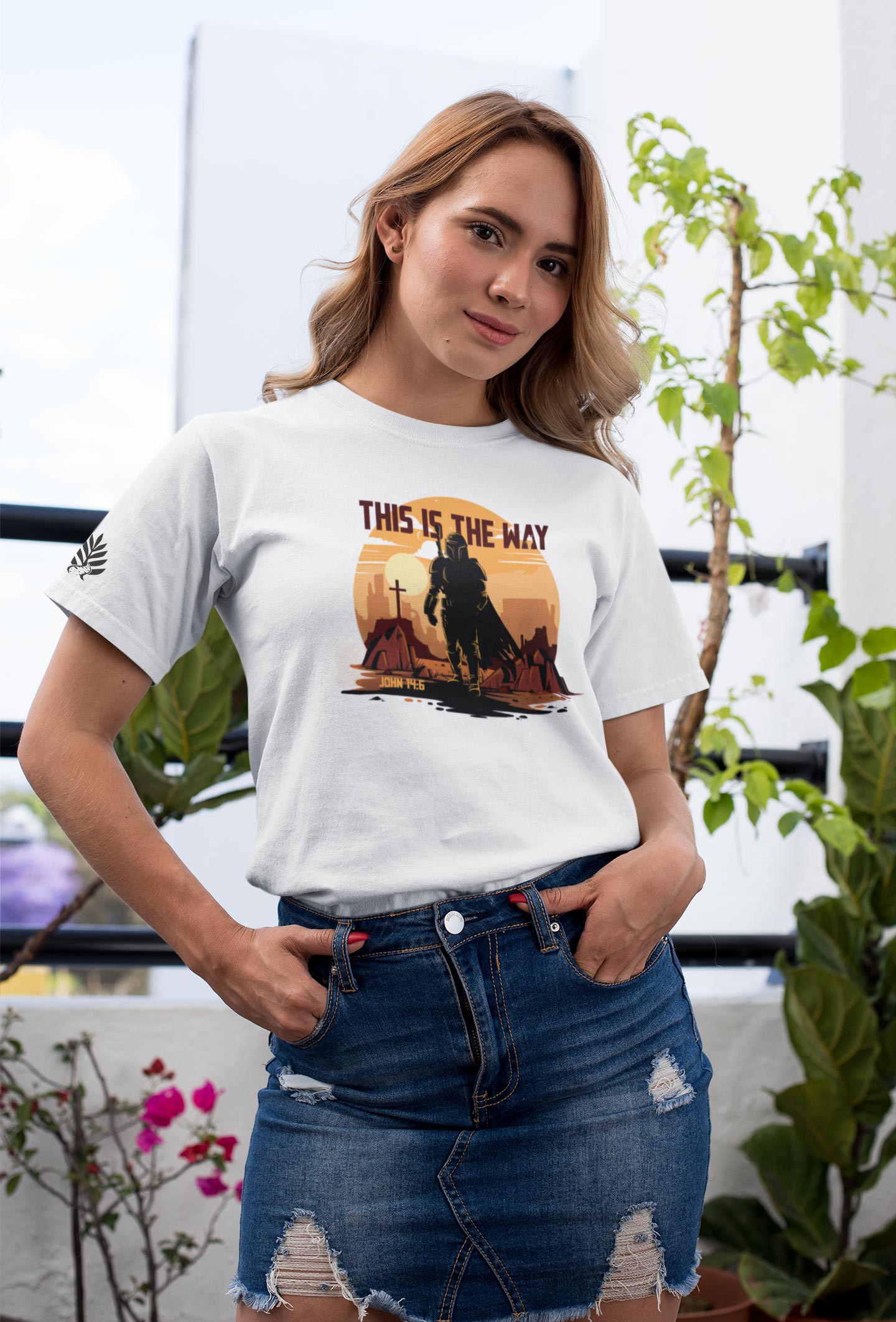 Woman wearing a white t-shirt with a graphic design and text, standing outdoors with plants in the background.