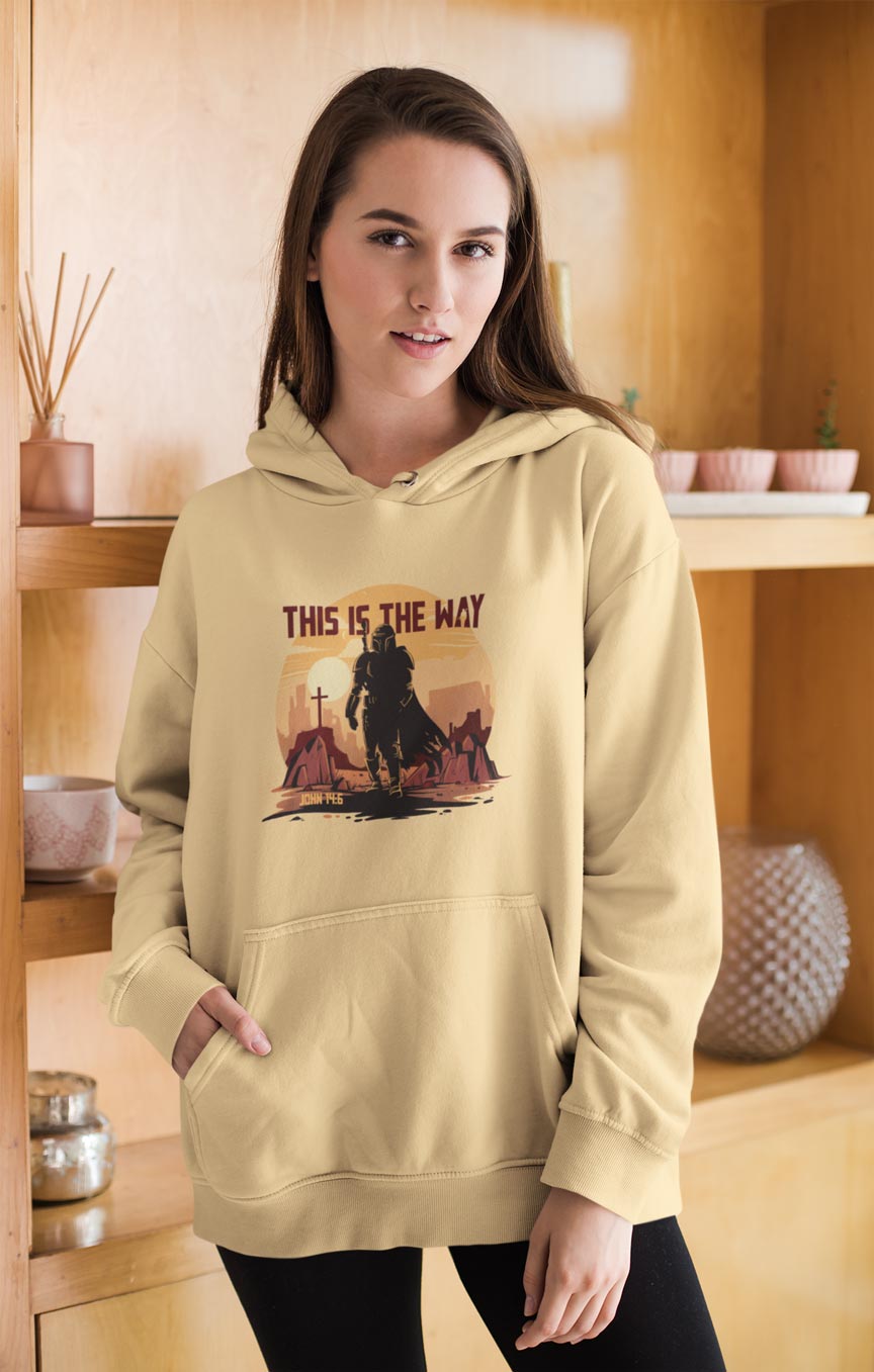 Woman wearing a beige hoodie with a graphic design and text in a room with wooden shelves.